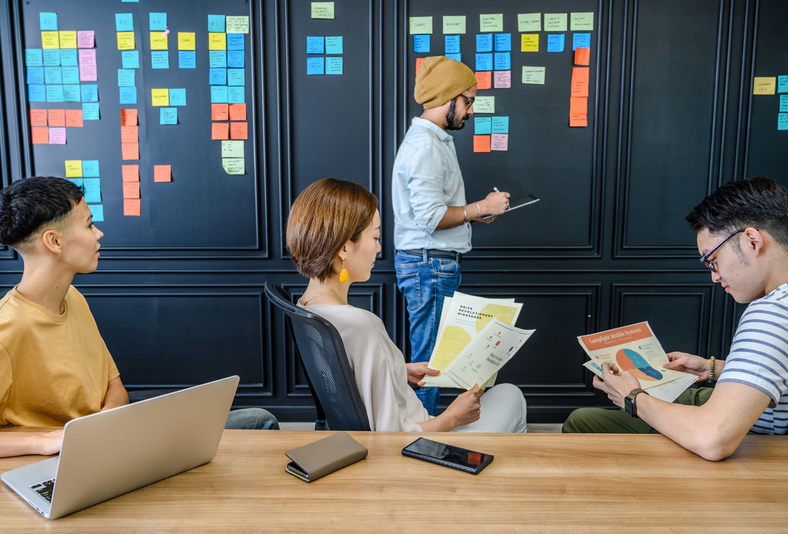 A diverse group reviewing plans, notes, and technology beside a wall organized with colorful requirements.